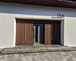 Voorkant van modern huis met WoodUpp verticale houten lamellen als gevelbekleding rondom de grijze voordeur; twee smalle ingebouwde buitenlampen in de lamellen, lange zwarte deurgreep en smalle glazen strook; nummer 13A boven; betonnen treden en straattegels op de voorgrond zichtbaar.