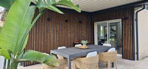 Terrasfoto met WoodUpp verticale houten lattenwand: donker gebrande lamellen gemonteerd in zwarte metalen kaders als gevelbekleding en hoekpaneel. Overdekt zitgedeelte met grijze tafel, rotan stoelen, sierplant en lichte tegelvloer. Zwarte vierkante wandlampen accentueren de strakke, warme WoodUpp-afwerking en houtnerf zichtbaar.
