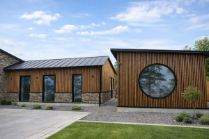 Een moderne woning met een buitenkant die is bekleed met WoodUpp producten. De gevels zijn voorzien van verticale houten lamellen, waarschijnlijk Thermo Es van WoodUpp, die een warme uitstraling geven. Het rechter gebouw heeft een opvallend groot rond raam, terwijl het linker gebouw een stenen basis heeft.