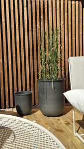 Close-up van een WoodUpp-achterwand van smalle verticale houten latten met zwarte tussenruimtes en een WoodUpp-terras van brede lichtgekleurde houten planken; daarnaast twee grijze bloempotten (een grote met hoog siergras, een kleinere leeg) en een deel van een wit loungestoelframe.