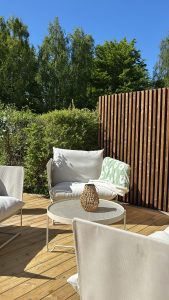 Zonnig terras met lichte lounge-stoelen en rond metalen tafeltje; op de achtergrond WoodUpp houten terrassenvloer met brede, naturel planken en knoesten, en een WoodUpp verticaal latten-scherm in warm bruin voor privacy. Sfeervolle rieten lantaarn en groen deken, omgeven door hoge bomen.