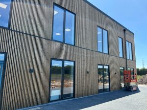 WoodUpp gevel: moderne tweelaagse gevelbekleding met smalle, verticale houten latten van WoodUpp, aangebracht rondom grote ramen met zwarte kozijnen. Donkere inbouwlampen en een mobiele hoogwerker staan voor de nieuw gebouwde kantoorgevel. Natuurlijke houtnerf en regelmatige ribbelstructuur zichtbaar. De gevel beslaat twee bouwlagen, met reflecties van blauwe lucht in het glas.