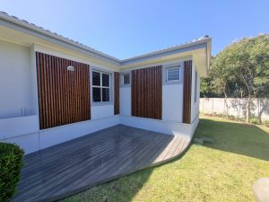 Hoek van een witte woning met WoodUpp donkerbruine houten lamellenpanelen langs de gevel en een verhoogd, grijs WoodUpp terras van smalle diagonale planken; dubbelglasraam en metalen buitenlamp, omliggend groen gazon, bomen en een heldere blauwe lucht op een zonnige dag.