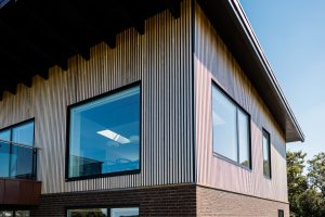 Hoek van modern huis bekleed met WoodUpp verticale houten lattengevel; slanke, lichtgekleurde houten lamellen zorgen voor ritme langs grote glazen ramen en balkon. WoodUpp-profielen vormen strakke, duurzame gevelbekleding boven donkere bakstenen plint, met overstekend dak en heldere lucht op de achtergrond.