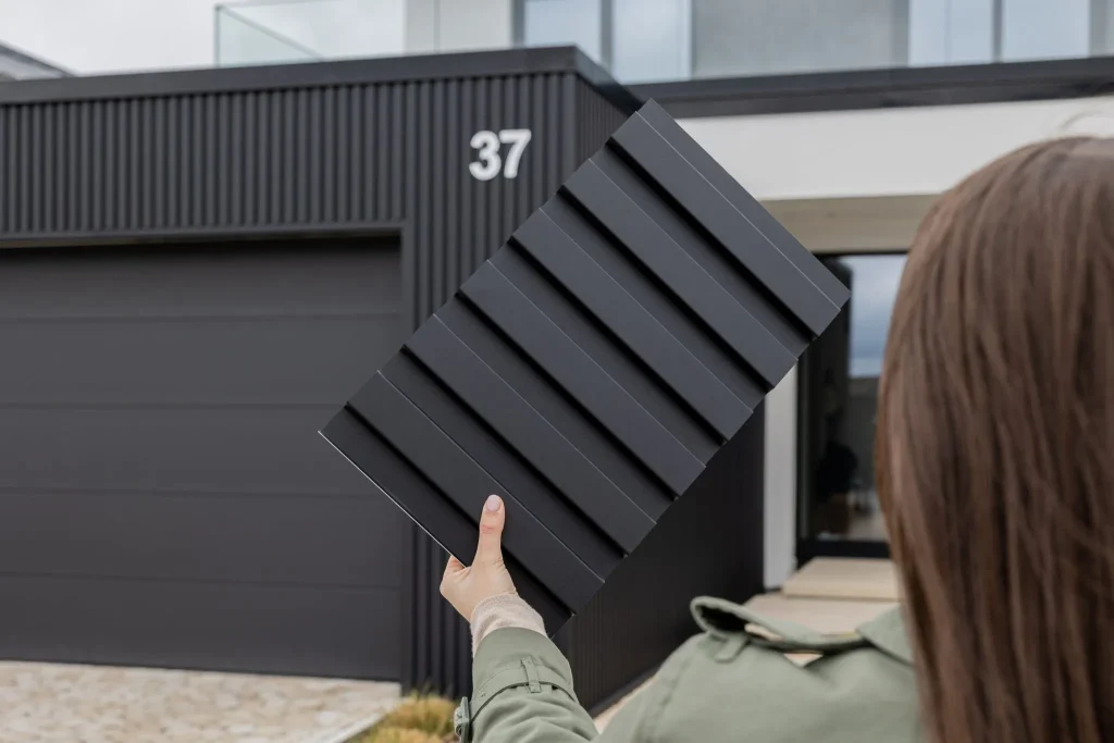 Persoon houdt een zwart WoodUpp monsterpaneel met brede, horizontale ribbels omhoog voor een modern huis. Op de achtergrond zijn WoodUpp buitengevelpanelen zichtbaar: verticale, matte zwarte latten en een bijpassende garagedeur. Duidelijke structuur, strak modern profiel en egale, duurzame donkere afwerking.