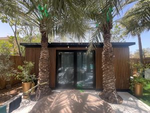 WoodUpp tuinkamer met verticale houten lattenpanelen als gevel- en schuttingbekleding, donkere aluminium kader en grote glazen schuifdeur; twee palmbomen en plantenpotten op betegeld terras creëren schaduw. Lattenpanelen tonen smalle, regelmatige houten stroken voor moderne, natuurlijke uitstraling. Sfeerverlichting hangt onder de zwarte dakrand en accentueert de warme bruine tint en houttextuur.