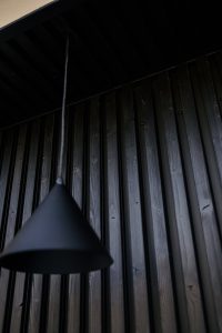 Close-up van een donker interieur met WoodUpp zwarte verticale houten lamellen als wandbekleding, zichtbaar in profiel met schroefgaten en houtnerf. Op de voorgrond hangt een matte zwarte conische hanglamp, deels onscherp, onder een zwart plafondpaneel, met nauwkeurig gefreesde groeven en donkere beits die textuur benadrukt.