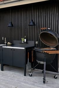 WoodUpp buitenruimte met zwarte WoodUpp buitenkeuken: metalen kast met geïntegreerde spoelbak, donker werkblad en klein wandplankje, bevestigd aan verticale zwarte WoodUpp houten wandbekleding. Rechts een open kamado-grill op wielen met houten zijplankjes en metalen schep, op lichtgrijze houten terrasplanken, sfeervol stijlvol.