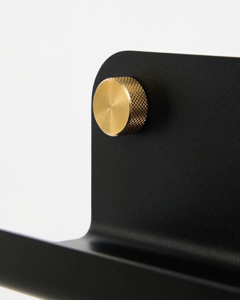 Close-up van een WoodUpp zwart matte metalen wandbeugel met afgeronde hoek, bevestigd door een goudkleurige, geribbelde messing knop. De foto toont het strakke, minimalistische ontwerp van WoodUpp: zwarte stalen plaat en contrasterende messing bevestigingsbout, met fijne textuur en zichtbare draaikrassen.