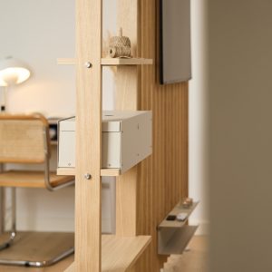 WoodUpp roomdivider van licht eiken met verticale latten en dunne planken, uitgerust met een lichtgrijze metalen opbergbox, een smal metalen plateau voor afstandsbedieningen en een klein plankje met een touwklosje; tegen het lattenpaneel hangt een televisie, met een bureau en rieten stoel op de achtergrond.