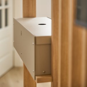 Close-up van een WoodUpp-installatie: beige metalen opbergbox met ronde kabeldoorvoer en klein ventilatieornament, gemonteerd tussen warme eikenhouten verticale latten van het WoodUpp-paneel. Strak, minimalistisch design met zichtbare schroeven en naadloze aansluiting tussen hout en metalen opbergunit voor kabels of accessoires, subtiel afgewerkt.