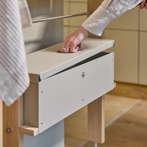 Close-up van een WoodUpp-opstelling: een compacte, matte witte, rechthoekige opbergbox met scharnierend deksel en ronde duimgreep, bevestigd aan een houten eiken frame onder een roede. Hand opent de metalen WoodUpp-box; zichtbare schroeven en geventileerd logo benadrukken het praktische, minimalistische ontwerp.