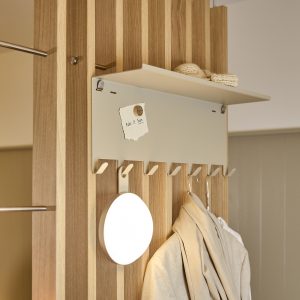 WoodUpp kapstokpaneel met verticale eiken latten, een smal bovenplankje, matte metalen achterplaat met magneet, zes beige metalen haken en ronde wandlamp/spiegel. Strak Scandinavisch ontwerp, functioneel en warm van kleur. Jas en sjaal hangen aan de haken, en een gebreide muts ligt op de plank.