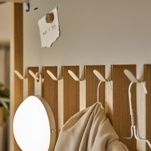 WoodUpp kapstokpaneel met verticale eiken latten en meerdere lichte metalen haken; aan de haken een metalen kledinghanger en een beige jas. Links een rond WoodUpp-wandlamp met zacht licht. Bovenaan een klein magnetisch houten knopje met een opgehangen briefje, in warme Scandinavische stijl.