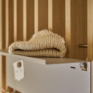 WoodUpp lattenwand van licht eiken met verticale latten en een ingebouwde, matte metalen brievenbus; bovenop het deksel ligt een opgevouwen crème, grofgebreid wollen sjaal. Rechts is een klein, rond metalen haakje zichtbaar, passend bij het minimalistische ontwerp met zachte, warme tonen.