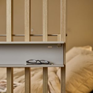WoodUpp houten room divider met verticale eiken latten, metalen bevestigingsstang en een smalle grijs-beige opbergplankje bevestigd aan de latten. Op het plankje liggen een bril en een klein boekje; gehard metalen haak en subtiele messing accentdetails zichtbaar in warme slaapkameromgeving.