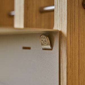 Close-up van een WoodUpp-eiken wandpaneel met bijbehorend WoodUpp metalen ophanghaakje/clip, goudkleurig met subtiele cirkelgravure. De metalen achterplaat en het haakje zijn bevestigd aan het hout; op de achtergrond vervaagde houten latten en een RVS-ophangstang zichtbaar, met matte, warme afwerking en zichtbare houtnerf.