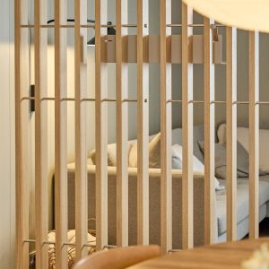 Close-up van een WoodUpp houten lamellenwand als roomdivider: licht eiken, smalle verticale latten verbonden met dunne metalen stangen, strak en zwevend ontwerp. Achter de lamellen zichtbaar een beige stoffen bank met kussens. Rustige, natuurlijke sfeer door warme houttinten en minimalistische afwerking.