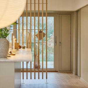 WoodUpp lamellenwand van licht eiken vormt subtiele ruimteverdeler vooraan in de keuken, met horizontale roestvrijstalen stangen en houten ophanghaken waaraan keukendoek, een netzakje en houten snijplankje hangen. De smalle, rechte lamellen accentueren het natuurlijke interieur en scheiden keuken van buitendeur, met elegante afwerking.