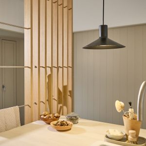 Rustige keukenhoek met WoodUpp lattenwand als roomdivider: licht eikenhouten verticale lamellen met ingebouwde metalen kledingrails en kleine haakjes. Op het aanrecht houten schaaltjes met noten, een glazen pot en keukenborstel; hanglamp boven het blad en neutrale grijze lambrisering op achtergrond.