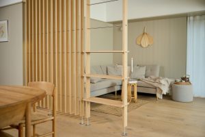 WoodUpp houten roomdivider met smalle verticale eiken lamellen en horizontale rvs-stangen, gecombineerd met een WoodUpp open ladderrek van eiken met planken en verstelbare metalen voetjes. Gezellige woonkameropstelling met beige bank, vloerkleed en houten meubels in warm, natuurlijk Scandinavisch interieur stijlvol.