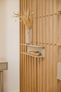 WoodUpp eiken lattenwand met smalle verticale lamellen naast een beige metalen WoodUpp wandplank met twee niveaus. Op de plank staan twee kleine houten schaaltjes en bovenop een glazen vaas met gedroogde bloemen, in een warme, minimalistische interieursetting. De latten tonen een natuurlijke houtnerf en de plank een subtiele matte afwerking.