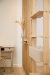 WoodUpp houten scheidingswand met verticale eiken lamellen, geïntegreerde open planken en een beige metalen opbergbox; aan de lamellen hangt een klein metalen wandplankje met een glazen vaas met droogbloemen en een houten schaaltje. Zicht op bureau en rieten stoel in zachte, neutrale tinten.