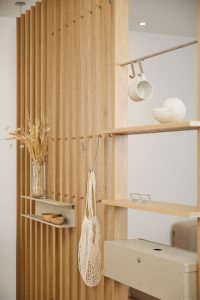 WoodUpp houten slatroomdivider met verticale eiken lamellen, geïntegreerde eiken planken, metalen haken en hangrail, en een beige metalen opbergbox plus smal wandplankje. Functionele, minimalistische en modulaire scheidingswand met open structuur en accessoires zoals kopje, vaas, netzak, kaarsjes en houten kommetjes, ideaal voor ruimteafbakening thuis.