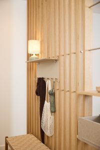 WoodUpp lamellenwand van licht eiken met een WoodUpp wandkapstok met plank en haakjes in beige; op de plank een lamp, aan de haken een donkerbruine sjaal, een katoenen netzak en opgerolde parapluhoes. Voor de wand een gevlochten zitbank en een zichtbare opberglade.