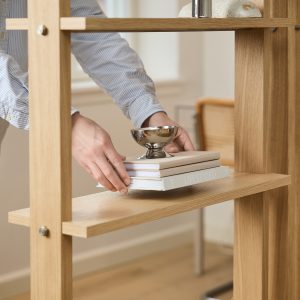 Hand plaatst een stapel boeken en een kleine zilverkleurige decoratieve schaal op een WoodUpp eikenhouten open kast. De WoodUpp-plank heeft slanke, lichte eiken schappen met zichtbare metalen boutverbindingen en een minimalistisch, modulair ontwerp dat ruimte en rust in de kamer benadrukt.