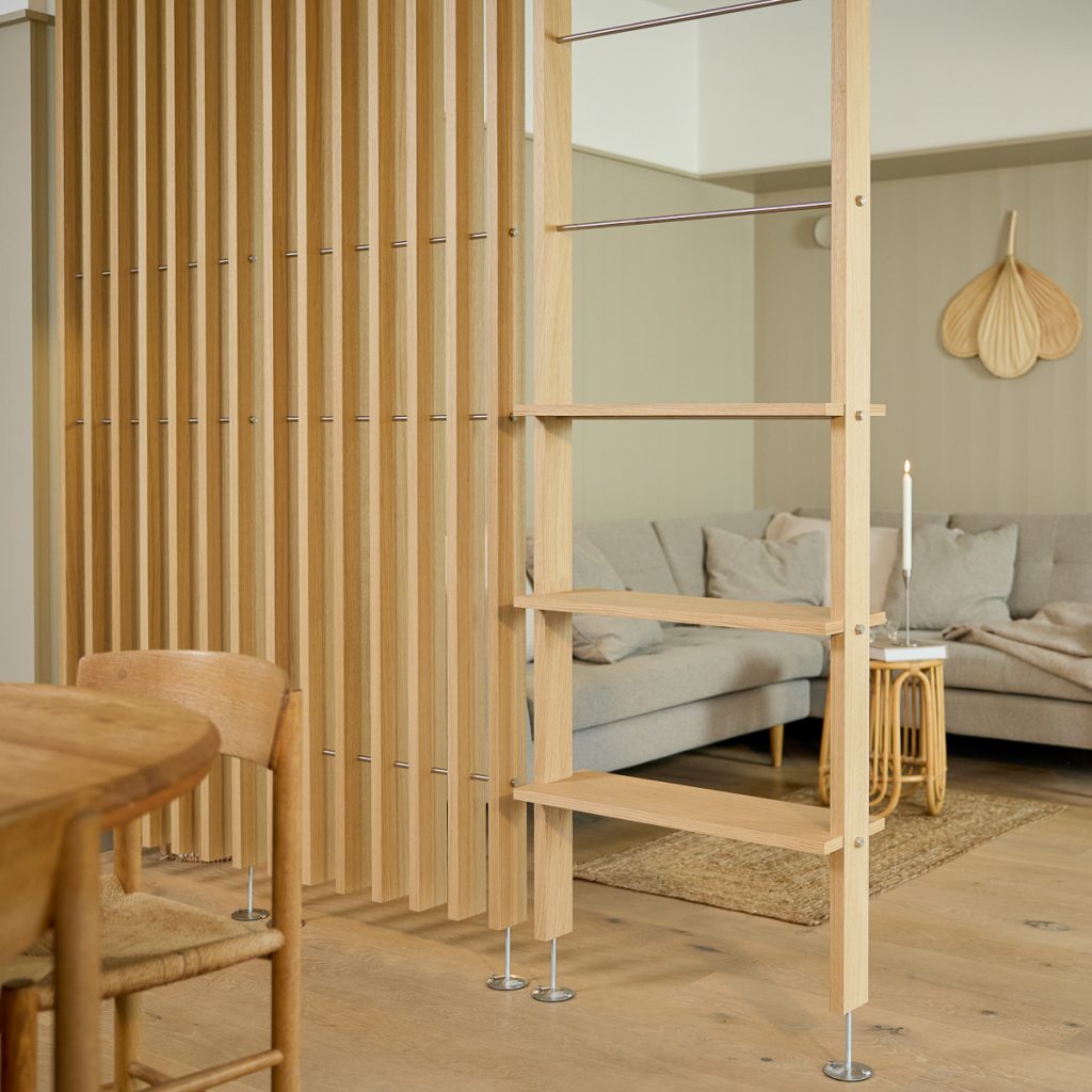 WoodUpp houten kamerverdeler en open rek: licht eiken lamellenwand met smalle verticale latten, metalen verbindingspennen en verstelbare voetjes, gecombineerd met een open plankensysteem van eikenplanken. Gezellige woonkamer op de achtergrond met grijze bank, rieten bijzettafeltje en brandende kaars voor een sfeervol natuurlijk interieur.