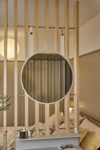 WoodUpp lattenwand in licht eiken vormt ruimteverdeler; aan metalen stangen hangt een WoodUpp ronde spiegel met witte rand, bevestigd met leren banden. Onder de spiegel een smalle WoodUpp wandplank met een mok, koptelefoon en een bril, uitzicht op bed met kussens en schemerlamp.