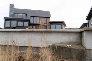 Moderne woning gezien achter een betonnen keermuur, met WoodUpp verticale houten latten als gevelbekleding en houten gevelprofielen rond ramen en balkon; grote zwarte glaspartijen, terras met plantenpotten en dor riet op de voorgrond en een laag modern bijgebouw met dezelfde latten bekleed.