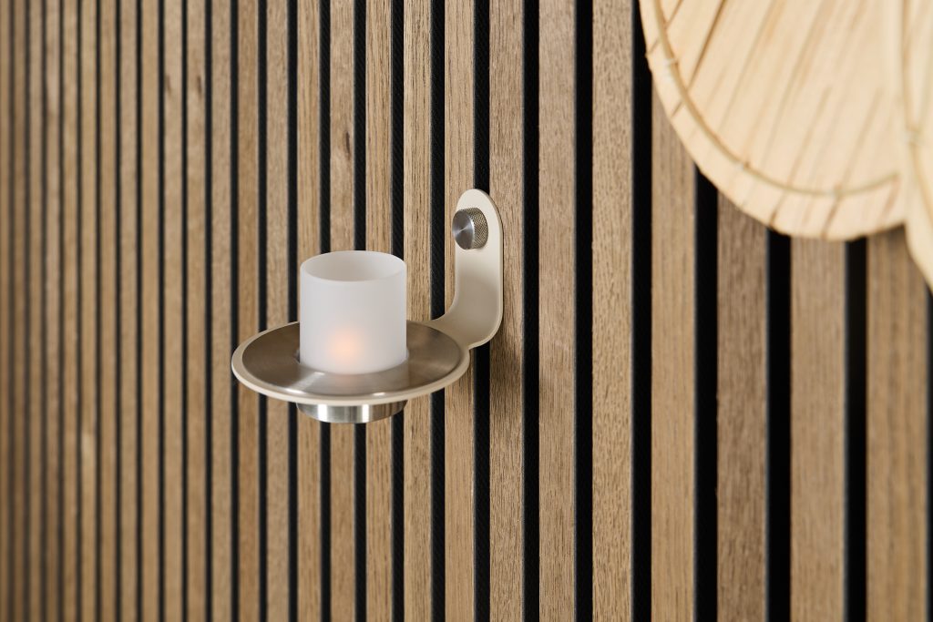 WoodUpp houten lamellenwand van smalle verticale eikenlatten met zwarte voegen en gelijkmatige tussenruimtes, gecombineerd met een WoodUpp metalen wandkandelaar in matte nikkelkleur met ronde schaal, cilindervormig mat glas en knopbevestiging, rechtsboven deels een rieten lampenkap zichtbaar voor extra warme sfeer.