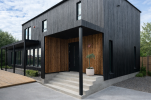 Een moderne woning met een zwarte houten gevel. De overdekte entree is opvallend bekleed met WoodUpp akoestische panelen. Deze verticale houten latten in een warme eiken kleur geven een stijlvol contrast en diepte aan de ingang bij de betonnen trappen en de donkere voordeur.